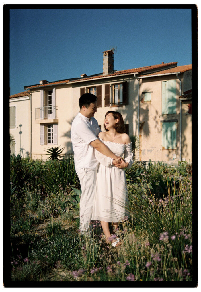 Couple laughing together during a honeymoon photo shoot in Antibes, standing in a garden with historic Riviera houses behind them